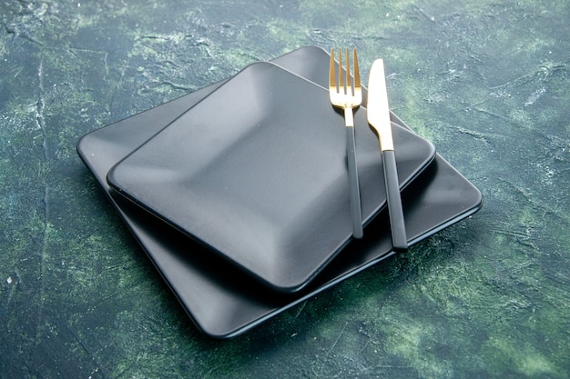vooraanzicht zwarte vierkante platen met gouden vork en mes op donkerblauwe achtergrond