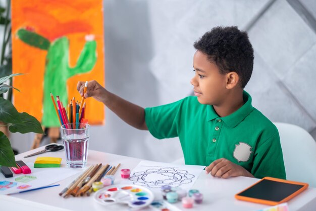 Voorbereiding, tekening. Profiel van een jongen met een donkere huidskleur in een groen t-shirt die een potlood uit glas haalt terwijl hij aan tafel zit en zich voorbereidt om bij daglicht te tekenen