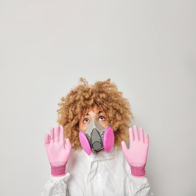 Voorzichtige vrouw houdt handpalmen richting camera om zichzelf te beschermen kwam in ecologische problemen heeft geen frisse lucht om te ademen draagt een wit beschermend pak gasmasker en handschoenen poseert binnenshuis kijkt erboven