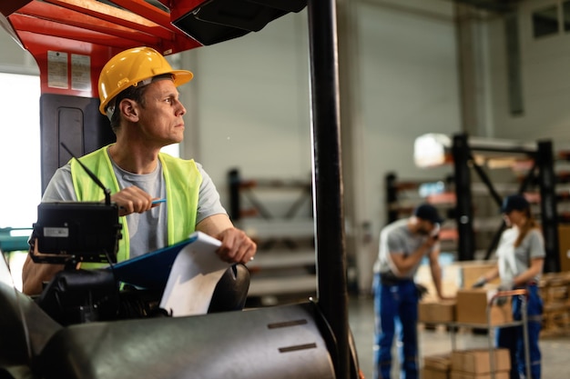 Vorkheftruckchauffeur die door papierwerk gaat en aan iets denkt terwijl hij in het distributiemagazijn werkt