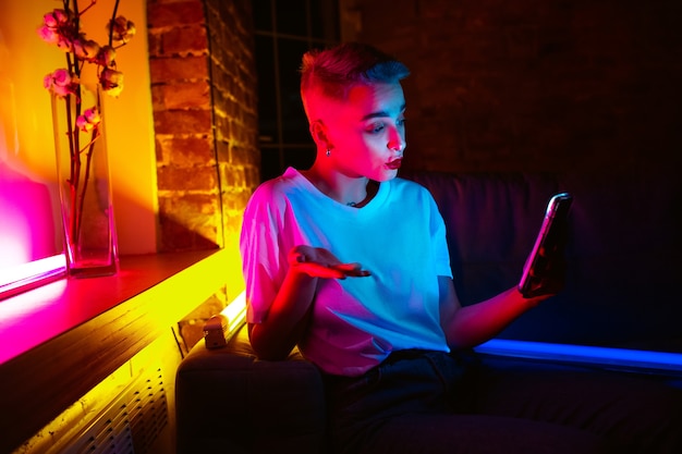 Vragen. Filmisch portret van stijlvolle vrouw in neon verlicht interieur. Afgezwakt als bioscoopeffecten, heldere neon-kleuren. Kaukasisch model met smartphone in kleurrijke lichten binnenshuis. Jeugd Cultuur.