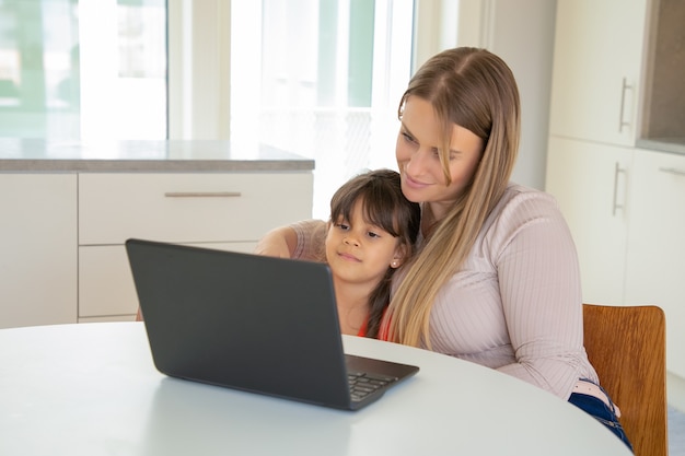 Vreedzaam meisje en haar moeder met behulp van laptop, aan tafel zitten en knuffelen, film kijken, display kijken.