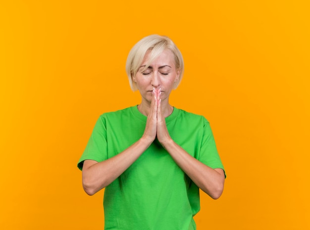 Vreedzame blonde Slavische vrouw van middelbare leeftijd die handen bij elkaar houdt bidden met gesloten ogen geïsoleerd op gele muur met exemplaarruimte