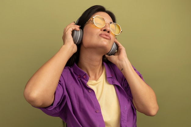 Vreedzame mooie brunette vrouw in zonnebril met koptelefoon staat met gesloten ogen geïsoleerd op olijfgroene muur