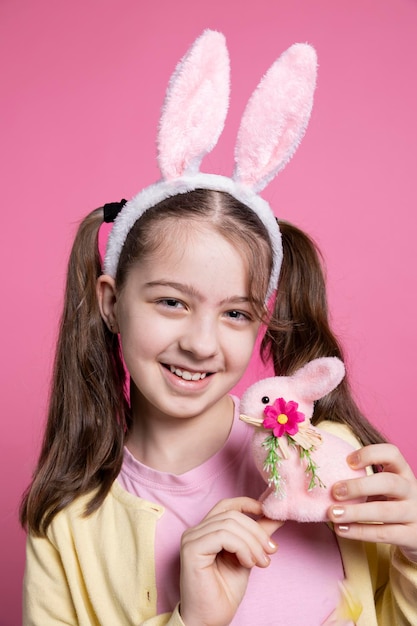 Gratis foto vreugdevolle mooie peuter met konijnenoren en pigtails die een roze konijn speeltje vasthoudt