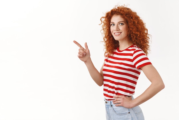 Vriendelijk behulpzaam charismatisch roodharige krullend jonge lange mooie vrouw pure schone huid wijzend linker bovenhoek staande halfgedraaide gestreepte tshirt hand taille zelfverzekerd draai camera glimlachen verzekerd