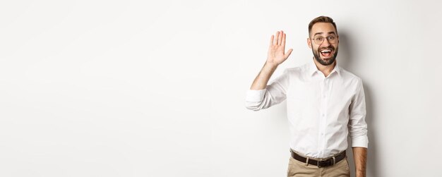 Gratis foto vriendelijk lachende man in glazen die hallo zegt zwaaiend hand in begroeting staande op witte achtergrond