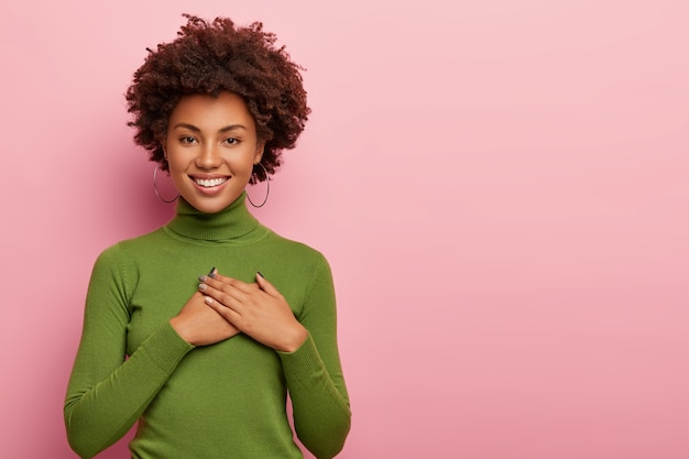 Gratis foto vriendelijk ogende donkere vrouw voelt zich dankbaar, spreekt dankbaarheid uit, heeft een hart gevuld met liefde, houdt beide handpalmen op de borst, draagt een groene coltrui, poseert op roze achtergrond, kopie ruimte