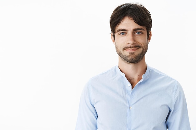 Vriendelijk ogende optimistische en knappe volwassen en mannelijke man met blauwe ogen en haren die vrolijk glimlachen terwijl hij terloops in blauw shirt tegen grijze muur staat