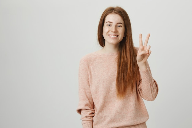 Vriendelijk roodharig meisje glimlachend en vredesteken tonen