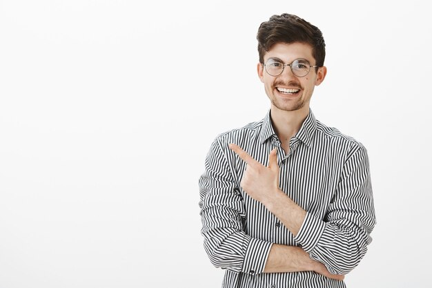 Vriendelijk, zorgeloos Kaukasisch vriendje met snor en baard in ronde bril, wijzend naar de linkerbovenhoek met vingerpistool en vrolijk glimlachend, vertellend over een geweldige plek om te zitten en koffie te drinken