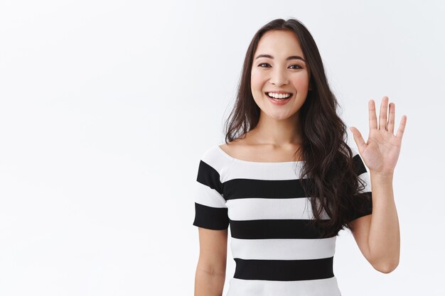 Vriendelijke, aangename en vrolijke Oost-Aziatische vrouw in gestreept t-shirt die palm opheft, hand groet, hallo of hallo zegt en lacht met vrolijke uitdrukking als gastvrije nieuwkomers, witte achtergrond