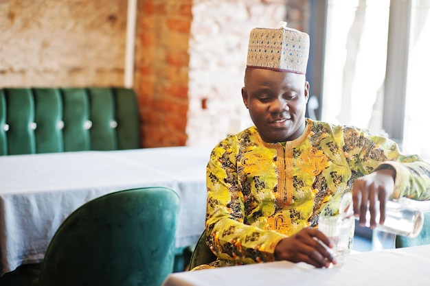 Vriendelijke afro man in traditionele kleding met pet zit in restaurant en giet water in een glas