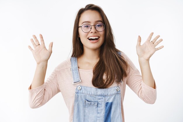 Vriendelijke en vrolijke jonge schattige vrouwelijke brunette in een bril en denim overall die beide handpalmen opheft, zwaaiend en hallo zeggend, groetend hallo gebaar en breed glimlachend naar elke gast over grijze muur.