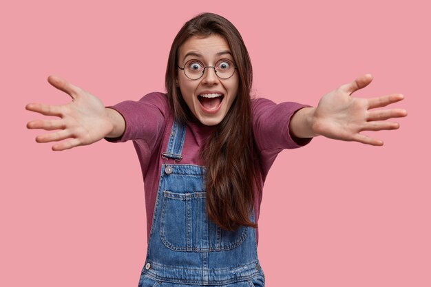 Vriendelijke gelukkige vrolijke vrouw geeft knuffel, gekleed in denim overall en paarse trui, kijkt met geluk