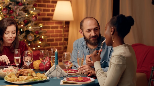 Vriendelijke jonge volwassenen die samen kletsen terwijl ze thuis genieten van het kerstdiner. Feestelijke familieleden die de wintervakantie vieren terwijl ze authentiek huisgemaakt eten eten.