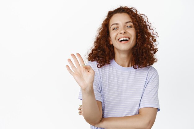 Vriendelijke jonge vrouw met rood krullend haar, zwaai met de hand, zeg hallo, groet iemand met opgeheven arm, vrolijk lachend, staande op een witte achtergrond