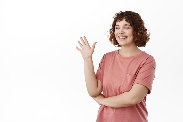 Vriendelijke jonge vrouw zwaaiend met de hand, opzij kijkend naar vriend en hallo zeggen, iemand groeten van sociale afstand, staande in t-shirt tegen witte achtergrond.