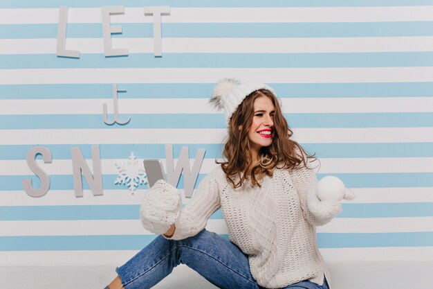 Vriendelijke, lieve jonge brunette kijkt verbaasd opzij en glimlacht oprecht. Indoor portret van meisje in witte kleren met telefoon