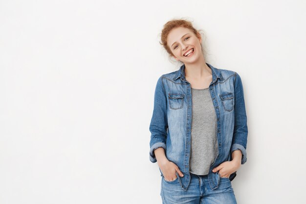 Vriendelijke positieve blanke roodharige vrouw met rommelig kapsel leunend op grijze ruimte met handen in de zakken