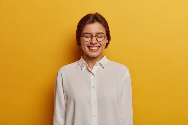 Vriendelijke positieve Europese studente kijkt met vrolijke uitdrukking, sluit ogen en grijnst naar camera, draagt ronde bril en wit overhemd