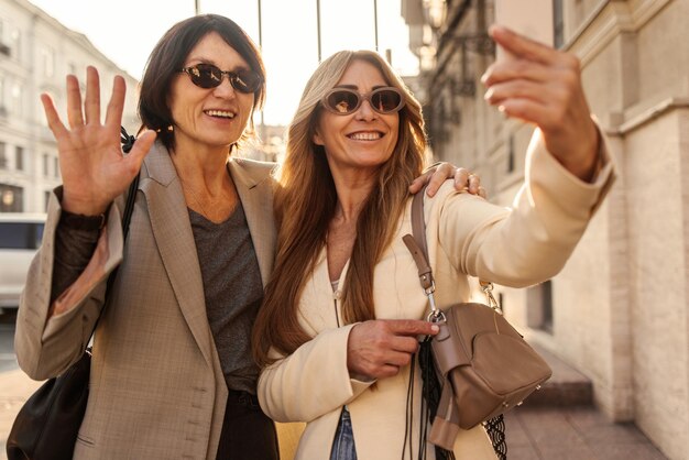 Vriendelijke volwassen dames met een lichte huid in zonnebrillen en jassen communiceren via videocommunicatie op straat Weekendplezierconcept