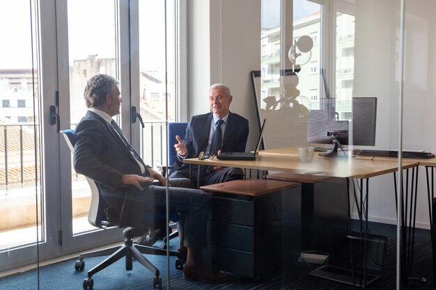 Vriendelijke volwassen zakenpartners bijeen in kantoor, zittend op de werkplek met laptop en praten. Bekijk door glazen wand. Portret bedrijfsconcept
