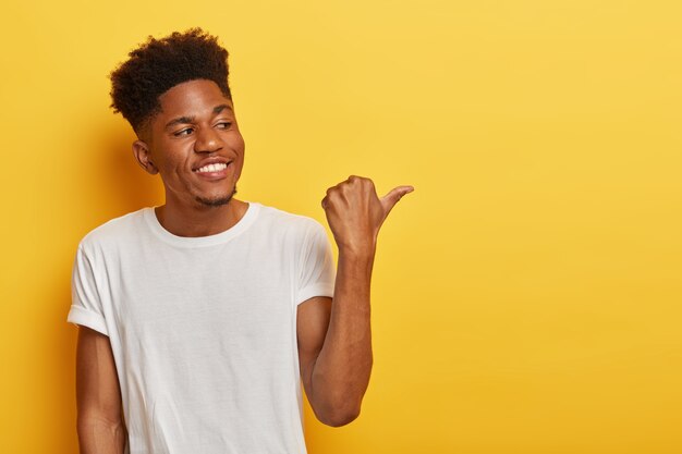 Vriendelijke vrolijke donkere hipster man bespreekt interessante kopie ruimte en wijst de duim weg aan de rechterkant, toont een plaats of richting, heeft een krullend trendy kapsel, draagt een casual wit t-shirt