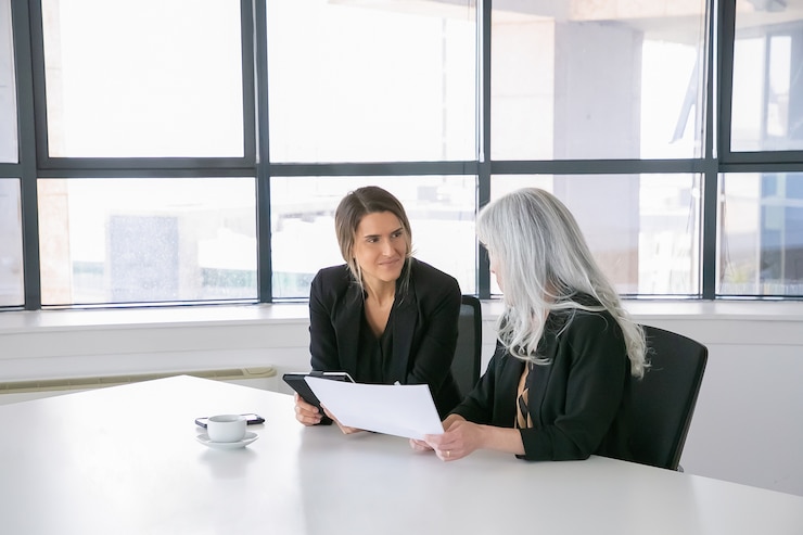 Gratis Foto vriendelijke vrouwelijke professionals die rapporten bespreken en analyseren. vrouwelijke ondernemers zitten samen, kijken naar documenten, tablet gebruiken en praten. communicatie concept