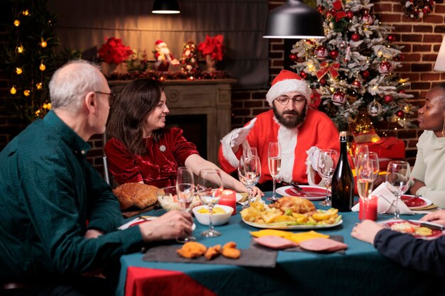 Vrienden die kerstmis vieren, een traditionele feestmaaltijd eten, een man die een kerstmankostuum draagt en een vrouw die aan het chatten is. Vakantie vieren, mensen verzamelen, mousserende wijn drinken