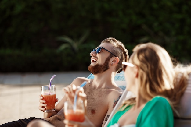 Vrienden glimlachen, cocktails drinken, liggend op ligstoelen bij zwembad