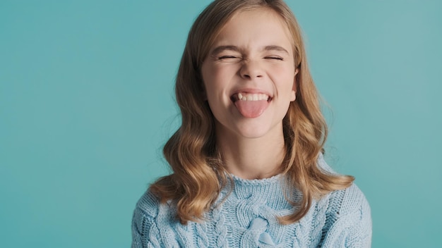 Vrij blond tienermeisje dat met golvend haar tong op camera toont die grappig over blauwe achtergrond kijkt