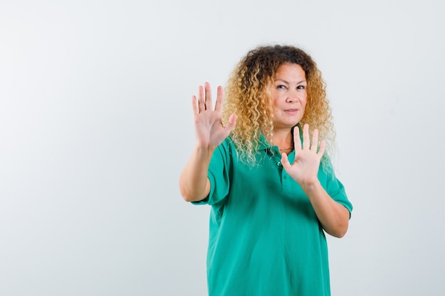 Vrij blonde vrouw die stopgebaar toont, handen houdt om zich in groen polot-shirt te verdedigen en bang kijkt. vooraanzicht.