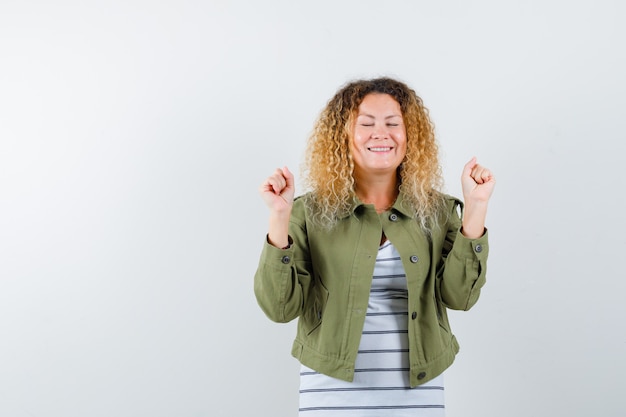 Vrij blonde vrouw die winnaargebaar in groen jasje toont en zalig kijkt. vooraanzicht.