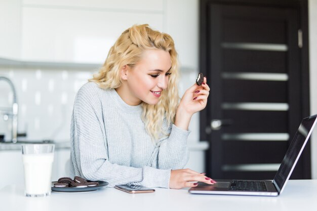 Vrij blonde vrouwenconsumptiemelk van een glas, die koekjes eten die laptop computer met behulp van terwijl het zitten bij de keukenlijst