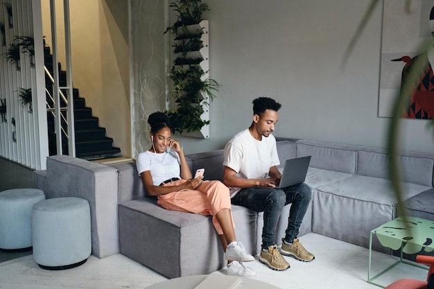 Vrij casual Afro-Amerikaanse vrouw die graag mobiele telefoon gebruikt terwijl jonge man in de buurt van werken met laptop op de bank in moderne co-working space