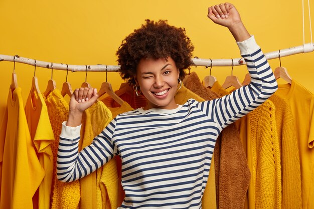 Vrij energieke Afro-Amerikaanse vrouw knipoogt, danst, draagt een striprd trui, staat tegen kledingroedes, koopt nieuwe kleding