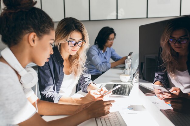 Vrij freelance copywriter sms-bericht op telefoon terwijl haar collega's op toetsenborden typen. Indoor portret van jonge programmeurs van verschillende etniciteiten die op kantoor werken.