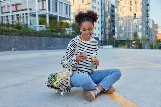 Vrij gelukkig tienermeisje met Afro-haar gebruikt mobiele telefoon om op internet te surfen zit gekruiste benen drinkt verfrissend water poses op weg op skateboard in stedelijke omgeving draagt nettas bladert internet