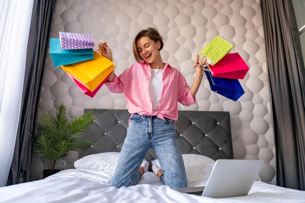 Vrij gelukkige vrouw die op bed springt die thuis online winkelt