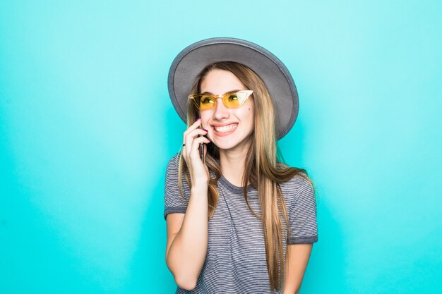 Vrij glimlachend jong model in maniert-shirt, hoed en transparante glazenbesprekingen op telefoon die op groene achtergrond wordt geïsoleerd