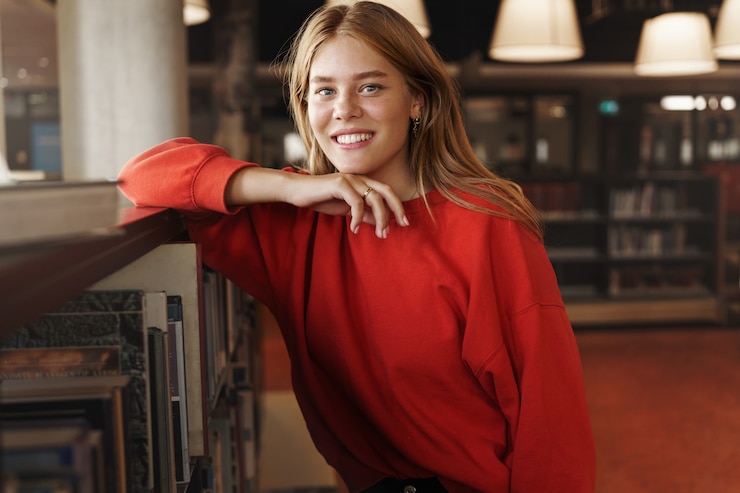 Gratis Foto vrij glimlachende roodharige vrouwelijke student, meisje dat op een boekenrek leunt.