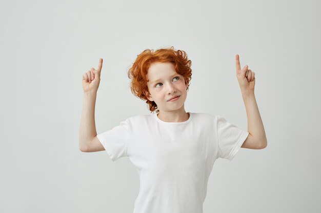 Vrij grappige kleine gemberjongen met sproeten die bovenkant met vingers op beide handen richt, licht glimlachend.