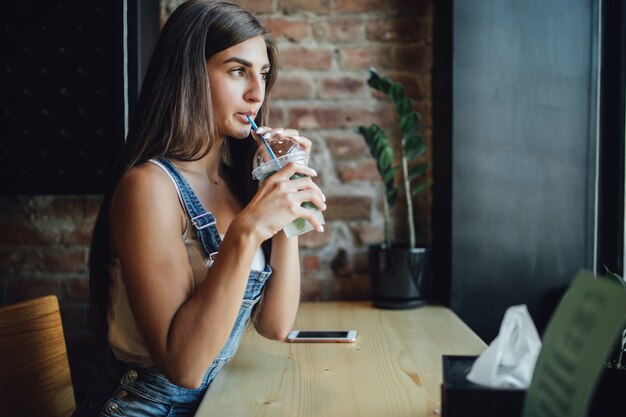 Vrij jong modelmeisje zit in het café voor het raam, werkt aan haar telefoon en drinkt een fris drankje