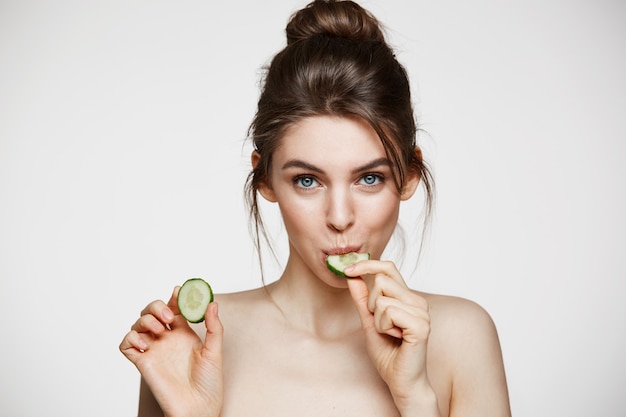 Vrij jong natuurlijk meisje dat met perfecte schone huid camera bekijkt die komkommerplak eet over witte achtergrond. Gezichtsbehandeling.
