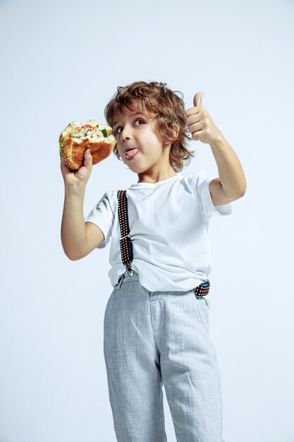 Vrij jonge krullende jongen in vrijetijdskleding op witte muur. Hamburger eten. Kaukasische mannelijke kleuter met heldere gezichtsemoties. Jeugd, expressie, plezier, fast food. Duim omhoog laten zien.