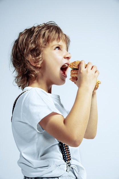 Vrij jonge krullende jongen in vrijetijdskleding op witte muur. Hamburger eten. Kaukasische mannelijke kleuter met heldere gezichtsemoties. Jeugd, expressie, plezier maken, fast food. Hongerig.