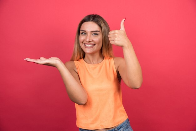 Vrij jonge vrouw die duimen op rode muur maken.