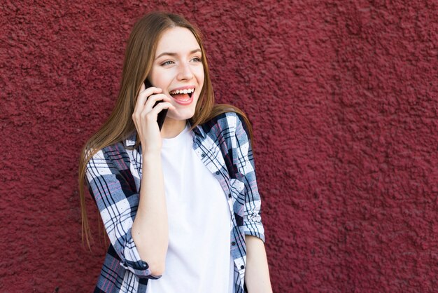 Vrij jonge vrouw die op cellphone met mond open tegen rode muur spreekt