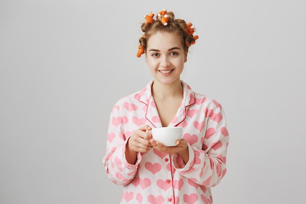 Vrij jonge vrouw in haarkrulspelden en pyjama's die ochtendkoffie drinken